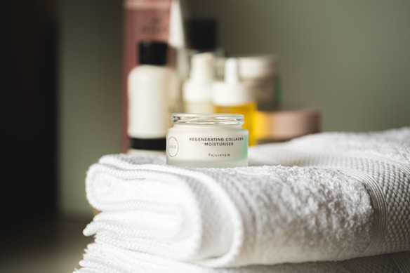 A small jar of regenerating collagen moisturizer is placed on a neatly folded, white towel. In the background, there are several blurred skincare bottles in various shapes and sizes.