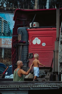 A bald man with a green sleeveless shirt holds the hand of a small child with blonde hair and shorts, standing on the back of a large red truck. An urban backdrop is visible with a building and billboard in the background.