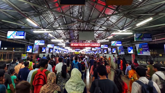 A crowded indoor railway platform with numerous people walking in different directions. Overhead, several digital screens displaying messages or advertisements are visible. A bright sign reads 'We Wish You A Happy Journey'. The platform is covered by a metal roof with visible lights and beams.