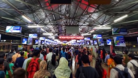 A crowded indoor railway platform with numerous people walking in different directions. Overhead, several digital screens displaying messages or advertisements are visible. A bright sign reads 'We Wish You A Happy Journey'. The platform is covered by a metal roof with visible lights and beams.