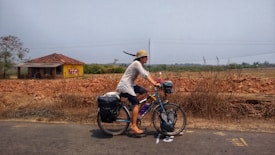 A person is riding a bicycle along a rural road with a load of luggage strapped on the back. The cyclist is wearing a hat and casual clothing. There is a small building with a tiled roof in the background, set against a dry, open landscape with sparse vegetation.