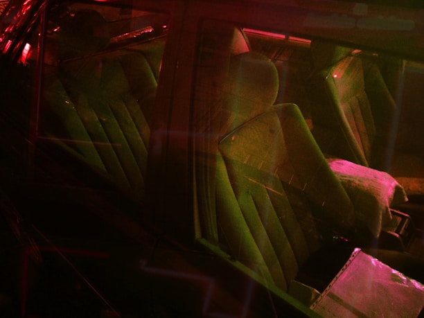The interior of a car illuminated by soft red and green lighting, highlighting the contours of the seats and the dashboard. The light creates a mysterious and somewhat futuristic ambiance.