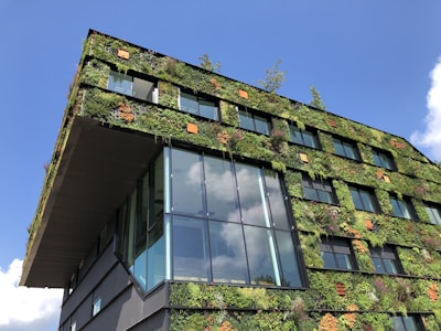 A modern building features green walls covered in various plants, creating a vertical garden effect. The structure has large reflective glass windows, and the vivid greenery contrasts with the blue sky in the background.