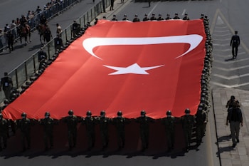A large group of uniformed individuals, likely soldiers, are holding and parading with a massive Turkish flag. The flag features a crescent and a star against a red background. The parade is taking place on a broad, barricaded road with spectators on one side. Soldiers are wearing helmets, and the scene suggests a national ceremonial occasion.