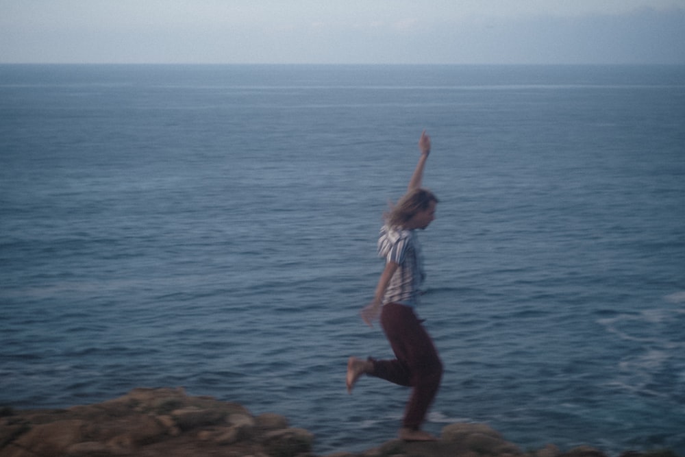 woman in white tank top and black pants standing on brown rock near body of water