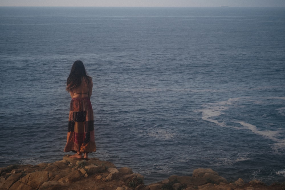 woman in red and black dress standing on brown rock near body of water during daytime