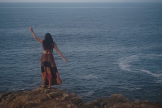 woman in red dress standing on rock formation near body of water during daytime