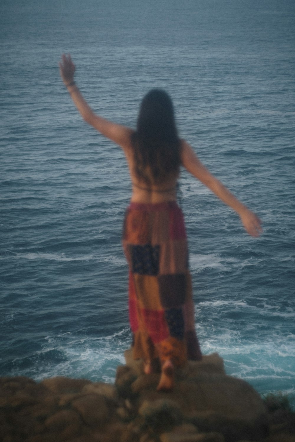 woman in red and black dress standing on rock near body of water during daytime