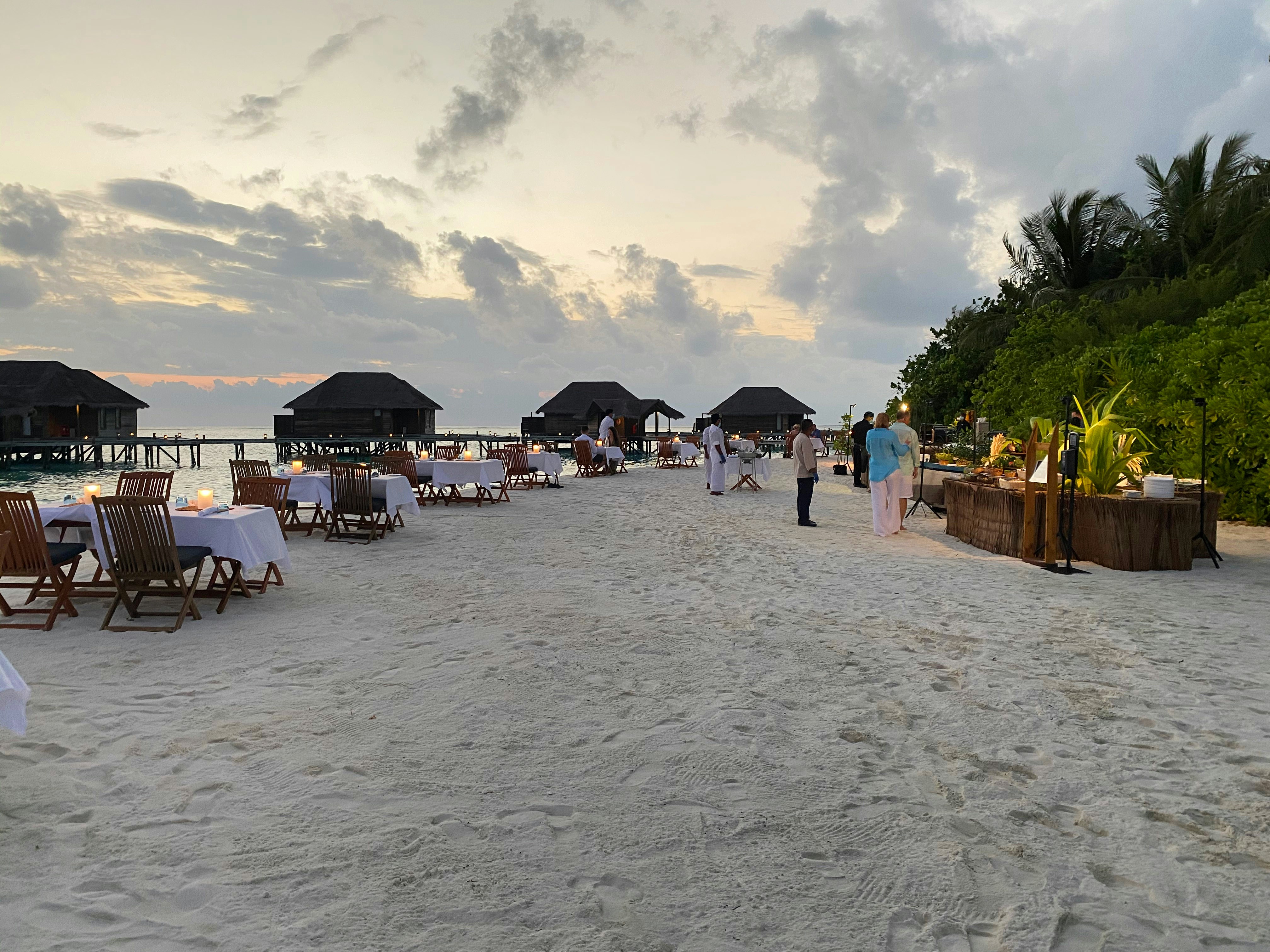 Elegant overwater dining table set at sunset with candles flickering, ocean stretching to horizon in warm orange and pink glow, fine dining presentation