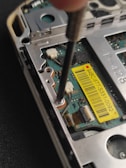 A close-up view of the inside of an electronic device, possibly a smartphone or a handheld gadget. The image focuses on a circuit board with small components, wires, and a yellow barcode sticker. A screwdriver is visible engaging with a screw, suggesting a repair or assembly process.
