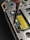 A close-up view of the inside of an electronic device, possibly a smartphone or a handheld gadget. The image focuses on a circuit board with small components, wires, and a yellow barcode sticker. A screwdriver is visible engaging with a screw, suggesting a repair or assembly process.