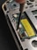 A close-up view of the inside of an electronic device, possibly a smartphone or a handheld gadget. The image focuses on a circuit board with small components, wires, and a yellow barcode sticker. A screwdriver is visible engaging with a screw, suggesting a repair or assembly process.