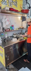 A street food vendor is preparing a dish at a small stainless steel food cart. The area is well-organized with various kitchen utensils and ingredients on the counter. A sign with Chinese characters is visible above the cart. The vendor is wearing a casual orange T-shirt, black pants, sandals, and a beige cap.