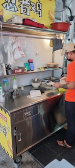 A street food vendor is preparing a dish at a small stainless steel food cart. The area is well-organized with various kitchen utensils and ingredients on the counter. A sign with Chinese characters is visible above the cart. The vendor is wearing a casual orange T-shirt, black pants, sandals, and a beige cap.