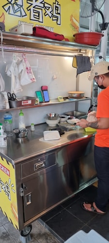 A street food vendor is preparing a dish at a small stainless steel food cart. The area is well-organized with various kitchen utensils and ingredients on the counter. A sign with Chinese characters is visible above the cart. The vendor is wearing a casual orange T-shirt, black pants, sandals, and a beige cap.