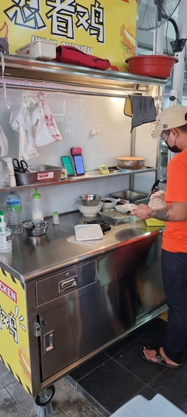 A street food vendor is preparing a dish at a small stainless steel food cart. The area is well-organized with various kitchen utensils and ingredients on the counter. A sign with Chinese characters is visible above the cart. The vendor is wearing a casual orange T-shirt, black pants, sandals, and a beige cap.