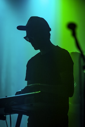 A silhouette of a person wearing a hat and sunglasses playing a keyboard, with a microphone on the side. The background is illuminated with vibrant shades of blue and green, creating a moody and atmospheric setting.