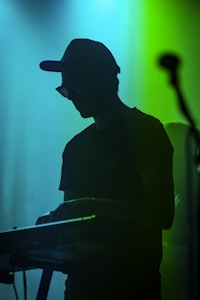A silhouette of a person wearing a hat and sunglasses playing a keyboard, with a microphone on the side. The background is illuminated with vibrant shades of blue and green, creating a moody and atmospheric setting.
