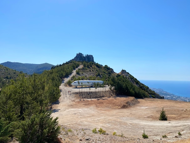 A secluded mountaintop location features a piece of an airplane surrounded by greenery and dirt paths, with sweeping views of the distant mountains and sea. Two small figures are walking near the aircraft, indicating the scale of the landscape.
