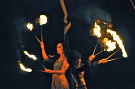 Two individuals are performing a fire dance. They are holding flaming sticks and manipulating the fire in a dark setting, creating patterns with the flames. One person is wearing a flowy outfit, while the other is dressed in darker attire. Smoke is emanating from the flames, adding to the dramatic atmosphere.