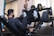 A person exercises on a leg press machine in a gym. The individual is wearing a black outfit and white sneakers, pushing against weights with their legs. The gym environment is equipped with various fitness machines and a poster on the wall.