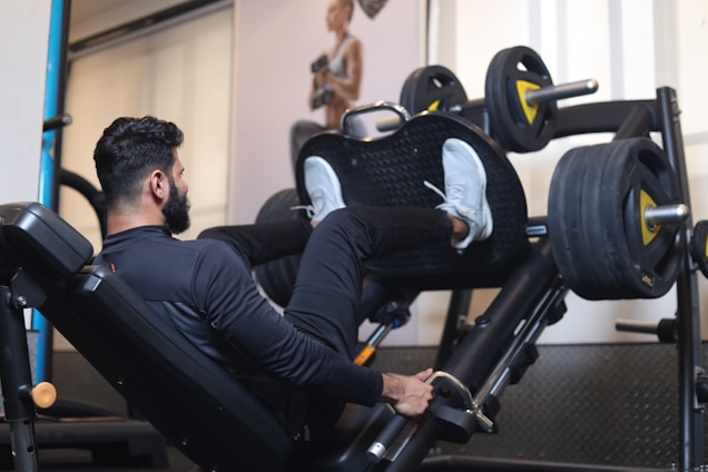 A person exercises on a leg press machine in a gym. The individual is wearing a black outfit and white sneakers, pushing against weights with their legs. The gym environment is equipped with various fitness machines and a poster on the wall.