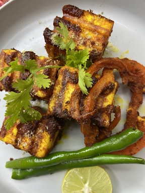 Grilled pieces of paneer with a charred, spiced coating are garnished with fresh coriander. Slices of grilled onion and two green chili peppers rest alongside a slice of lime, all arranged on a light-colored plate.