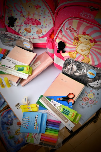 The image contains two children's backpacks with cartoon sheep designs, surrounded by various school supplies. The supplies include notebooks, scissors, a ruler, highlighters, an eraser, and colored markers. The backpacks have pink and red tones with decorative elements.