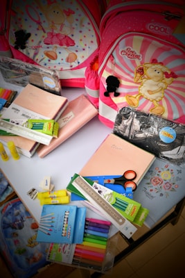 The image contains two children's backpacks with cartoon sheep designs, surrounded by various school supplies. The supplies include notebooks, scissors, a ruler, highlighters, an eraser, and colored markers. The backpacks have pink and red tones with decorative elements.