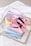 Several brightly colored rectangular cases, including pink, blue, and yellow, are stacked on a reflective tray. Each case contains makeup items like lipsticks and brushes. There are also decorative rings and a gold necklace nearby, placed on a textured white surface.