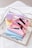 Several brightly colored rectangular cases, including pink, blue, and yellow, are stacked on a reflective tray. Each case contains makeup items like lipsticks and brushes. There are also decorative rings and a gold necklace nearby, placed on a textured white surface.