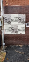 A brick wall displays repeated posters with the phrase 'KEEP KIDS OUT OF CAGES' overlaid on a pattern resembling chain-link fencing. There is a utility pole next to the wall, and some weathering is visible on the posters and the wall.