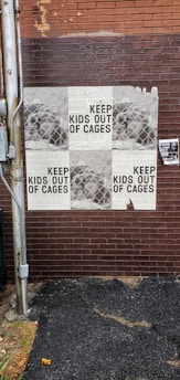 A brick wall displays repeated posters with the phrase 'KEEP KIDS OUT OF CAGES' overlaid on a pattern resembling chain-link fencing. There is a utility pole next to the wall, and some weathering is visible on the posters and the wall.