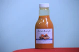 A bottle filled with an orange-colored drink labeled 'Asam Kunyit Minuman Sehat' placed on a red surface with a plain blue background. The label includes images of turmeric and other ingredients.