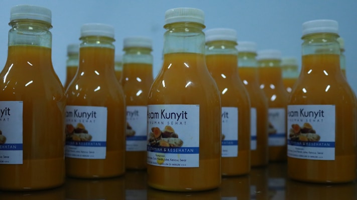 Several bottles filled with a yellow liquid are arranged on a reflective surface. Each bottle is sealed with a white cap and has a label featuring text and images. The liquid inside appears to be a turmeric-based drink.