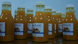 Several bottles filled with a yellow liquid are arranged on a reflective surface. Each bottle is sealed with a white cap and has a label featuring text and images. The liquid inside appears to be a turmeric-based drink.
