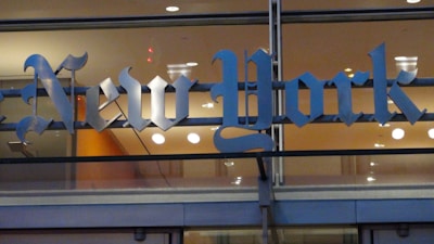 An elegant, metallic sign with the text 'New York' in a traditional Gothic font. The letters are mounted on a glass wall with warm interior lighting and reflections visible through the glass.
