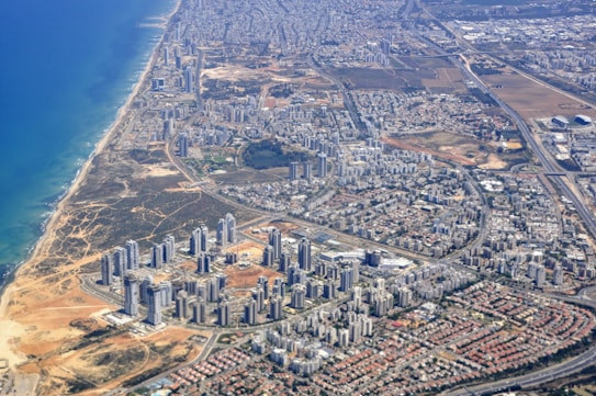High-rise buildings and dense urban development near a coastline. There are sandy areas near the sea moving towards a vast expanse of residential and commercial buildings. Roads and infrastructure are visible, connecting different parts of the city. Beyond the developed area, there are patches of open land and industrial zones.