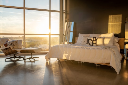 A modern bedroom with large floor-to-ceiling windows allowing sunlight to pour in, creating a warm and inviting atmosphere. A neatly made bed with several decorative pillows sits beside a stylish chaise lounge, with a small side table and a tall mirror adding to the decor.