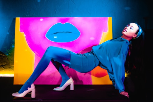 A person is posing in front of a vibrant and colorful artwork featuring a large set of lips. The background is a mix of pink and yellow, creating a striking contrast. The person is dressed in blue attire, lying back dramatically with legs extended and wearing white high-heeled shoes. The lighting and bold colors contribute to a dramatic and artistic composition.