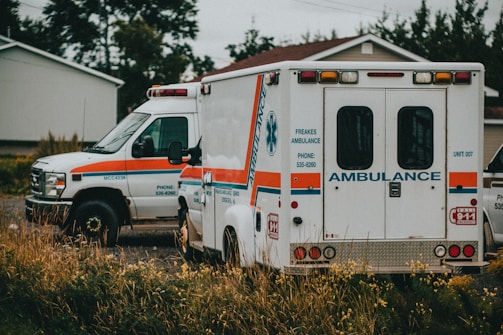 Two ambulances are parked, one facing forward and the other showing its rear side. They are situated in a grassy or overgrown area near a residential house. The vehicles are white with orange and blue stripes.