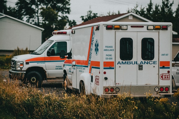 Two ambulances are parked, one facing forward and the other showing its rear side. They are situated in a grassy or overgrown area near a residential house. The vehicles are white with orange and blue stripes.