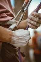 A close-up view of a person's hands, covered in chalk, gripping a climbing rope attached to a harness. The hand is secured to a carabiner, showcasing the preparation for rock climbing or a similar sport. The background is blurred, focusing on the hands and equipment.