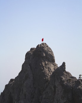 A rugged mountain peak topped with a small, vibrant red flag against a clear blue sky. The rocky surface is textured and appears steep, casting shadows that highlight the structure of the rock.