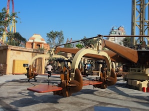 A thrilling amusement park ride with seats and harnesses suspended from a central rotating structure. The setting includes various architectural structures resembling ancient buildings and lush greenery. In the background, a colorful roller coaster can be seen.
