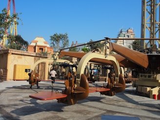 A thrilling amusement park ride with seats and harnesses suspended from a central rotating structure. The setting includes various architectural structures resembling ancient buildings and lush greenery. In the background, a colorful roller coaster can be seen.