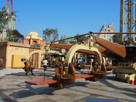 A thrilling amusement park ride with seats and harnesses suspended from a central rotating structure. The setting includes various architectural structures resembling ancient buildings and lush greenery. In the background, a colorful roller coaster can be seen.
