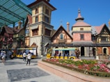 A whimsical, fairy-tale styled village scene featuring colorful half-timbered buildings with stone and brick elements, steep roofs, and decorative chimneys. Bright yellow trolley-like vehicles are parked in the foreground. People can be seen walking around the paved square, some taking photos and exploring. A vibrant flowerbed with various colored flowers sits in front of the buildings, adding a lively touch to the scene.
