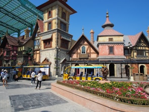 A whimsical, fairy-tale styled village scene featuring colorful half-timbered buildings with stone and brick elements, steep roofs, and decorative chimneys. Bright yellow trolley-like vehicles are parked in the foreground. People can be seen walking around the paved square, some taking photos and exploring. A vibrant flowerbed with various colored flowers sits in front of the buildings, adding a lively touch to the scene.