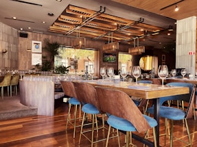 A well-decorated modern restaurant interior featuring wooden elements and ample natural light. The room is furnished with elegant wooden chairs with blue cushions, matching large tables set with fine glassware and napkins. The ceiling has stylish hanging light fixtures and the walls are adorned with plants and decorative art pieces. The environment exudes a combination of sophistication and warmth.